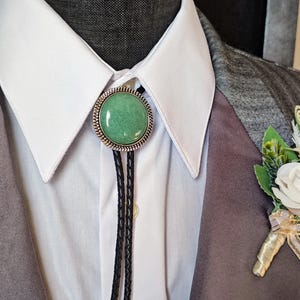 Puede incluir: Un bolo tie con una piedra preciosa verde en un engaste plateado. El bolo tie se lleva sobre una camisa blanca de cuello con una chaqueta de traje gris.