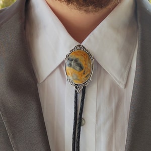 May include: A silver bolo tie with a yellow and gray stone set in a decorative frame. The bolo tie is worn with a white collared shirt and a gray suit jacket.