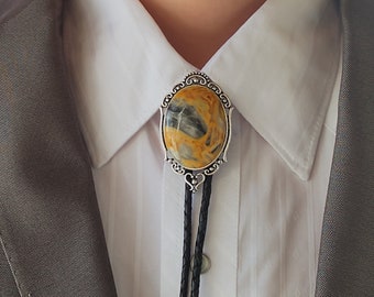 May include: A silver bolo tie with a yellow and gray stone set in a decorative frame. The bolo tie is worn with a white collared shirt and a gray suit jacket.