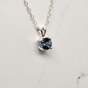 May include: A silver necklace with a round, deep blue gemstone pendant. The pendant is set in a four-prong setting and hangs from a delicate chain. The gemstone appears to be approximately 0.25 inches in diameter.