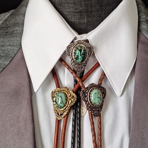 May include: Three bolo ties with turquoise-colored stones and decorative metalwork. The bolo ties have brown, black, and tan leather cords. The ties are displayed on a white collared shirt and gray blazer.