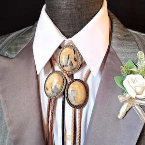 Può includere: Un bolo tie in pelle marrone, nero e beige con tre ciondoli ovali in pietra. Le pietre sono di colore marrone chiaro con vortici bianchi e grigi. La cravatta è indossata su una camicia bianca con colletto e una giacca di abito grigia.