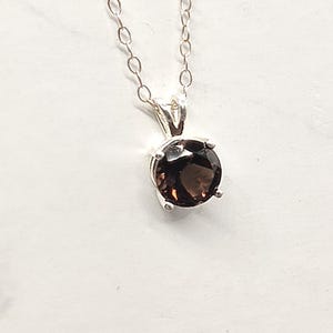 May include: A silver necklace featuring a round, dark brown gemstone pendant. The pendant is set in a four-prong setting and hangs from a delicate silver chain. The gemstone has a polished, reflective surface, and the chain is composed of small, interconnected links.