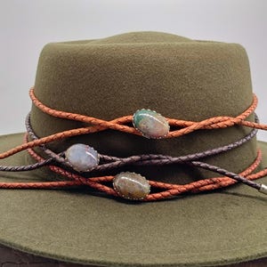 Op de afbeelding: Een olijfgroene hoed met drie gevlochten leren banden in bruin en oranje. De banden zijn versierd met drie ovale edelstenen in zilveren zettingen. De hoed heeft een klassieke fedora-vorm.