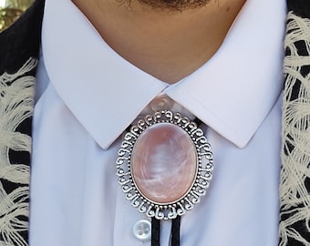 May include: A silver bolo tie with an oval, light pink stone center and a decorative border. The tie features a black braided cord and is worn over a white collared shirt.