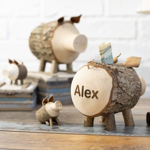 May include: Three wooden piggy banks in the shape of pigs. The largest piggy bank has the name "Alex" carved into it. The piggy banks are made from tree bark and have a rustic look.