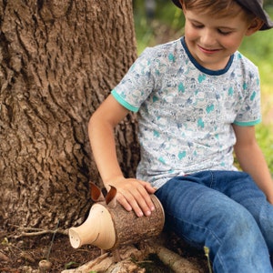 May include: A young person wearing a blue and white patterned shirt sits next to a tree trunk. They are holding a wooden piggy bank shaped like a pig with brown ears and a snout. The piggy bank is made to look like tree bark.