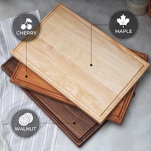 Puede incluir: Tres tablas de cortar de madera apiladas una encima de la otra. La tabla superior es de arce, la tabla del medio es de cerezo y la tabla inferior es de nogal.