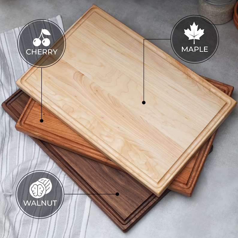 Puede incluir: Tres tablas de cortar de madera apiladas una encima de la otra. La tabla superior es de arce, la tabla del medio es de cerezo y la tabla inferior es de nogal.