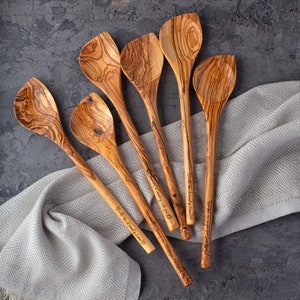 May include: Six wooden spoons with different engraved sayings. The spoons are made of olive wood and have a natural brown color. The spoons are arranged in a fan shape on a white and gray patterned fabric.