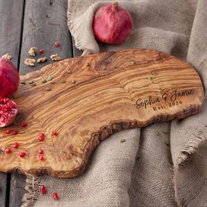 Op de afbeelding: Een houten serveerplank met een natuurlijke rand, gegraveerd met "Sophia & Jamie Est. 2026". De plank is omgeven door granaatappels, zaden en walnoten. De plank is gemaakt van olijfhout.