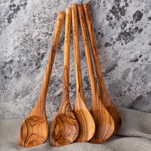 May include: Five wooden spoons with engraved messages. The spoons are made of olive wood and have a natural brown color. The messages on the spoons are: "Welcome in scooping with you", "All you Knead is Love", "You are my recipe for happiness", "All you Knead is Love", and "You are my recipe for happiness".
