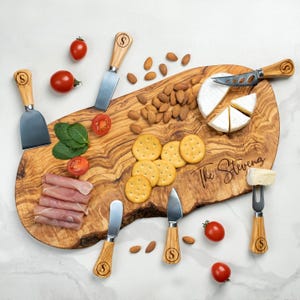 Op de afbeelding: Een olijfhouten serveerplank met de gegraveerde tekst "The Stevens" is omringd door kaasmesjes, crackers, amandelen, vleeswaren, tomaten en kaas. De messen hebben houten handvatten met de letter "S".