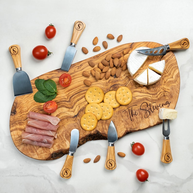 Op de afbeelding: Een olijfhouten serveerplank met de gegraveerde tekst "The Stevens" is omringd door kaasmesjes, crackers, amandelen, vleeswaren, tomaten en kaas. De messen hebben houten handvatten met de letter "S".