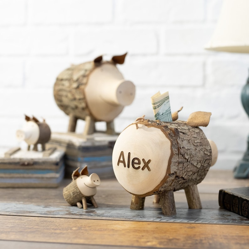 May include: Three wooden piggy banks in the shape of pigs. The largest piggy bank has the name "Alex" carved into it. The piggy banks are made from tree bark and have a rustic look.