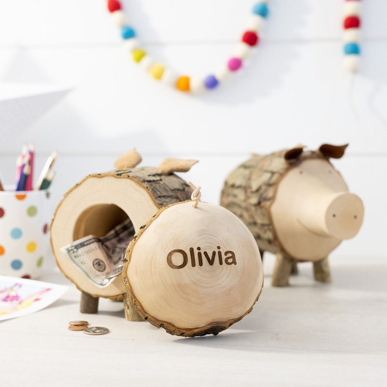 May include: Two wooden piggy banks shaped like pigs, one with a removable lid, with the name "Olivia" engraved on the lid. The piggy banks are made from natural wood with bark still attached.