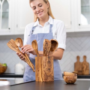 May include: A set of wooden kitchen utensils in a wooden holder. The utensils include a spoon, a spatula, and a ladle. The holder is made of olive wood and has a natural wood grain.