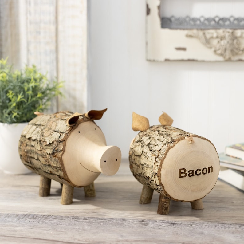 K&ouml;nnte beinhalten: Zwei h&ouml;lzerne Sparschweine in Form von Schweinen, eines mit dem Wort "Bacon" in die Seite geschnitzt. Die Sparschweine sind aus Baumst&auml;mmen gefertigt und haben kleine Holzbeine.