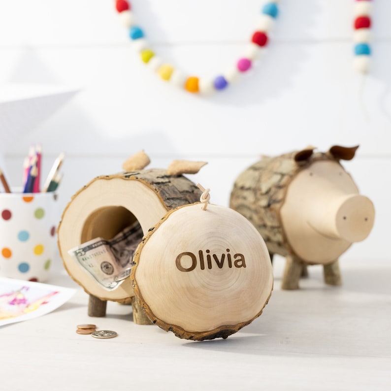 K&ouml;nnte beinhalten: Ein h&ouml;lzernes Sparschwein in Form eines Schweins mit einem Schlitz f&uuml;r M&uuml;nzen. Das Sparschwein ist aus einem Baumstamm gefertigt und hat den Namen "Olivia" auf der Seite eingraviert.