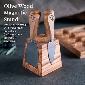 May include: Olive wood cheese knife stand with two magnetically held knives. The stand is a square block with a beveled top. The knives have wooden handles with the letter "M" engraved on them.