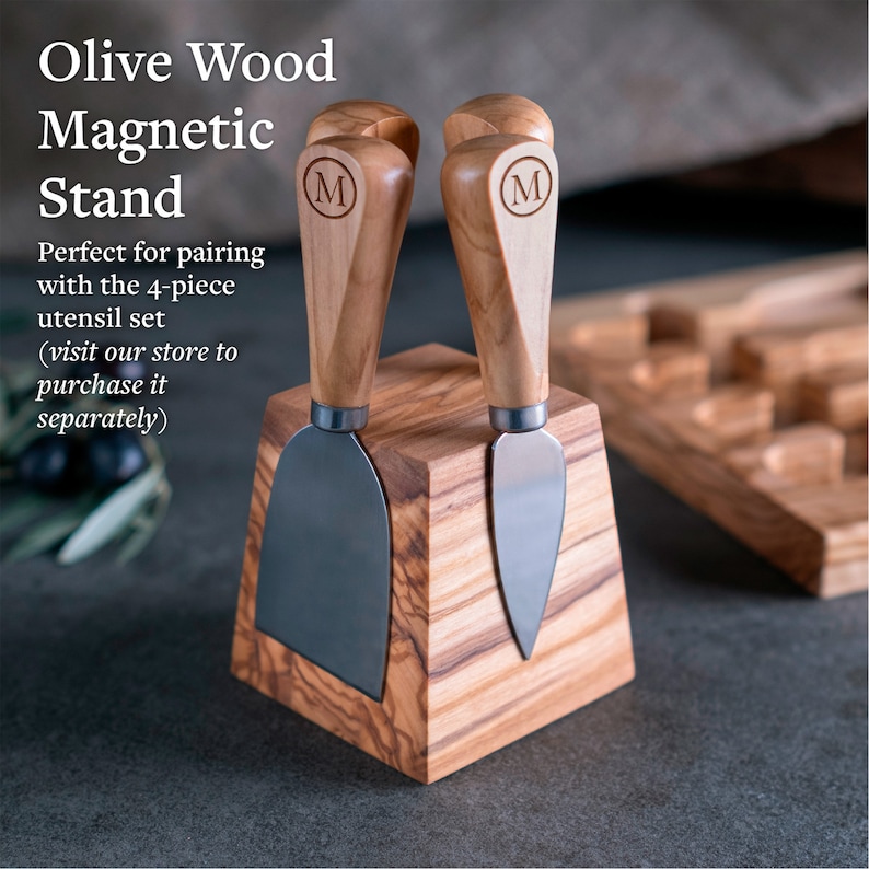 May include: Olive wood cheese knife stand with two magnetically held knives. The stand is a square block with a beveled top. The knives have wooden handles with the letter "M" engraved on them.