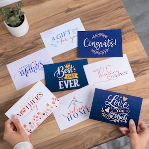 May include: A collection of greeting cards with various messages, including "Congrats!", "Best Dad Ever", and "Love You So Much". The cards feature floral and geometric designs in blue, white, and red, arranged on a wooden surface.
