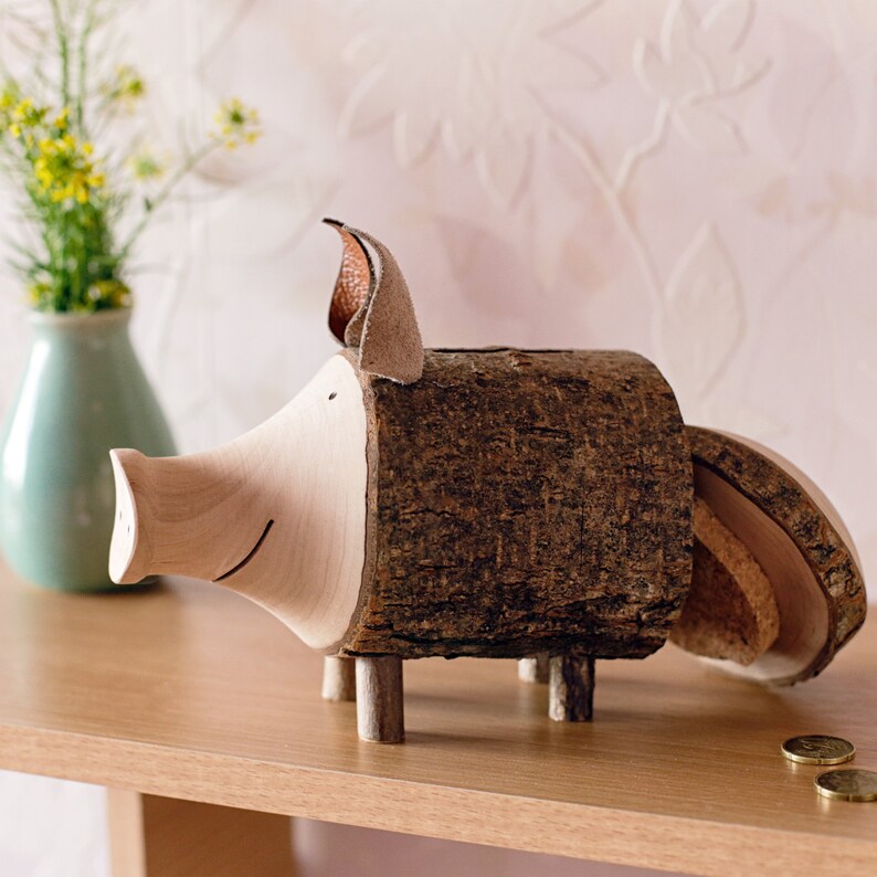 May include: A wooden piggy bank shaped like a pig with a brown bark exterior and a light wood snout and ears. The piggy bank has a slot in the back for coins and a removable bottom for access to the coins.