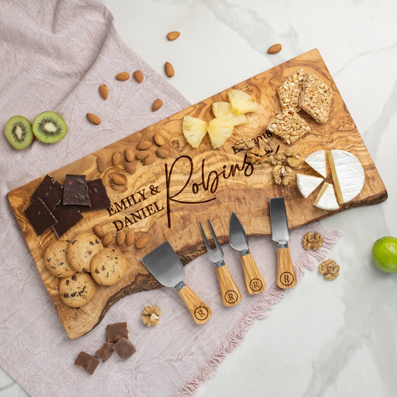 K&ouml;nnte beinhalten: Servierbrett aus Olivenholz mit einer nat&uuml;rlichen Kante, graviert mit den Namen "Emily & Daniel Robins" und dem Datum "Est. 2018". Das Brett ist mit verschiedenen Snacks gef&uuml;llt, darunter K&auml;se, Cracker, Obst, N&uuml;sse und Schokolade. Es enth&auml;lt auch vier K&auml;semesser mit Holzgriffen.