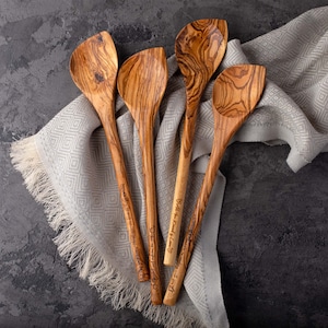 May include: Set of four olive wood spoons with engraved messages. The spoons are on a light gray and white patterned fabric.