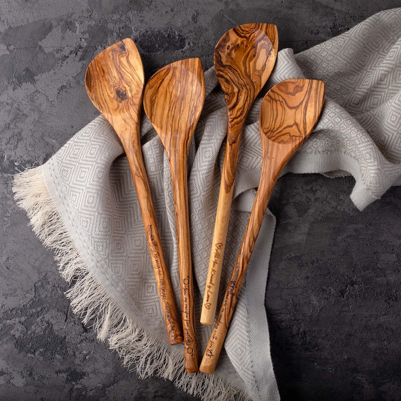 May include: Set of four olive wood spoons with engraved messages. The spoons are on a light gray and white patterned fabric.
