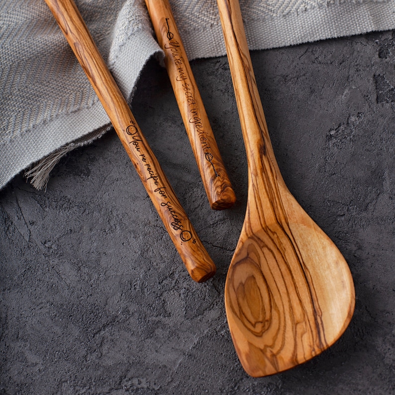 May include: Three wooden spoons with engraved sayings. The spoons are made of olive wood and have a natural wood grain. The sayings on the spoons are "You're my secret ingredient", "You're the recipe for success", and "You're my secret ingredient".