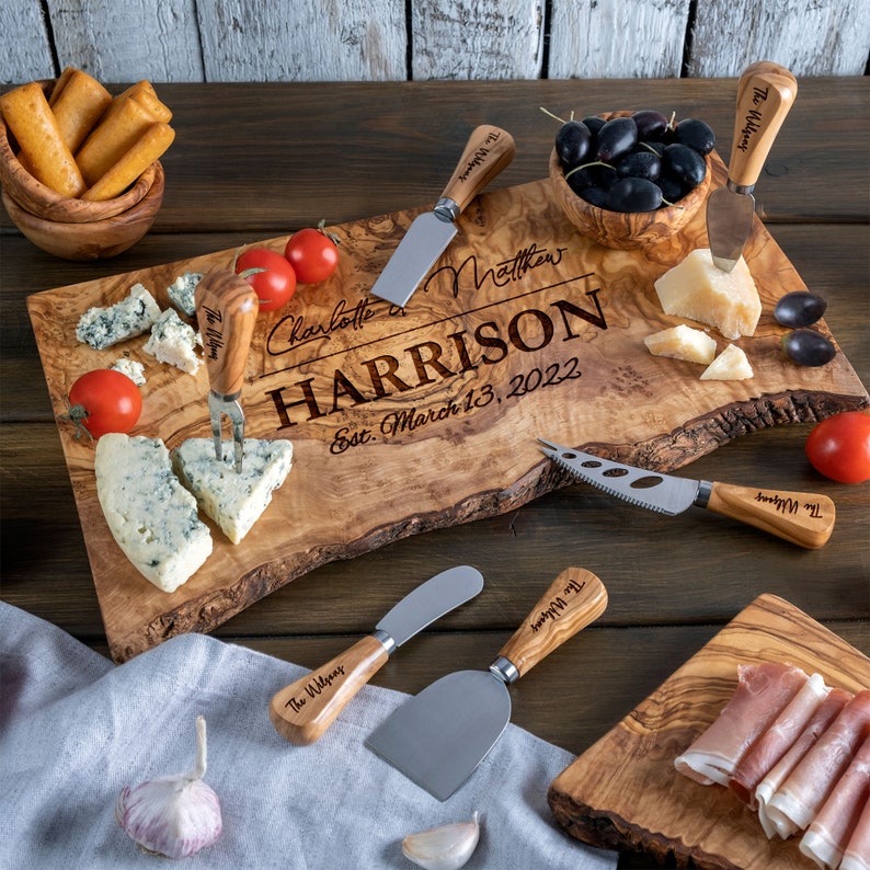 K&ouml;nnte beinhalten: Ein Holzbrett zum Schneiden mit einer personalisierten Inschrift, die "Charlotte & Matthew HARRISON Est. March 13, 2022" lautet. Das Brett ist von K&auml;se, Oliven, Trauben und anderen Lebensmitteln umgeben. Es gibt auch mehrere kleine Holz-K&auml;semesser mit der Aufschrift "The Wilsons" auf den Griffen.