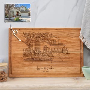 Puede incluir: Una tabla de cortar de madera personalizada con una ilustración grabada con láser de una casa y el texto "Lucas & Sophia Est. 2023".