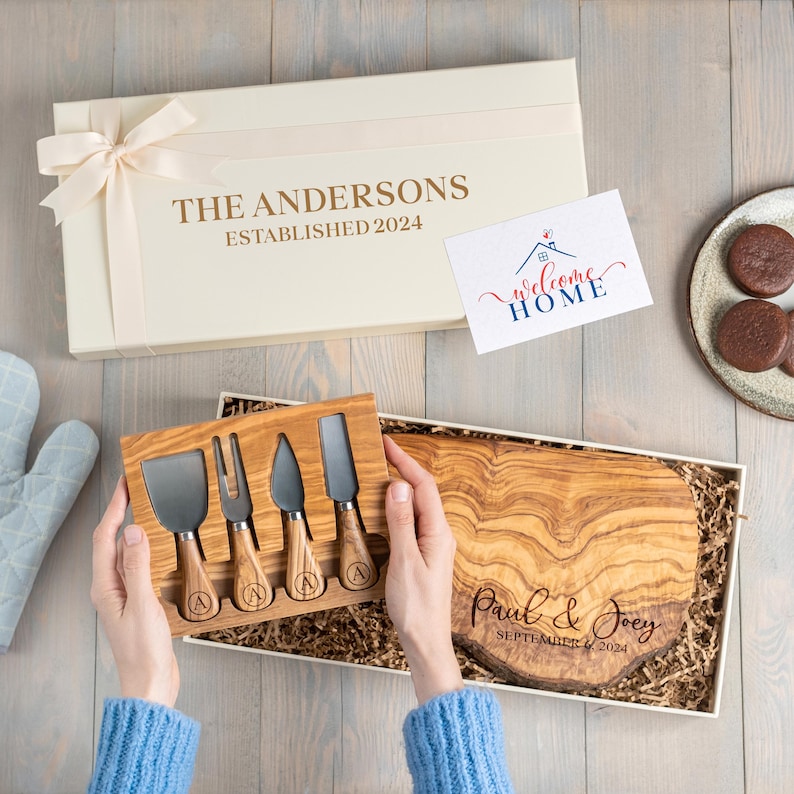Op de afbeelding: Een cr&egrave;mekleurige geschenkdoos met strik, met een houten kaasplank en kaasmesjes. De kaasplank is gegraveerd met "Paul & Joey SEPTEMBER 6, 2024". Een kaart leest "Welcome Home".