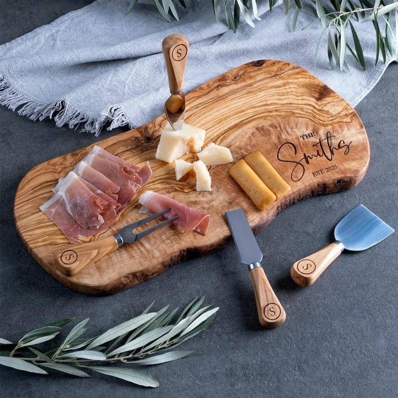 Op de afbeelding: Een houten kaasplank met kazen, vleeswaren en crackers. De plank bevat een kaassnijder, vork en spreader. De plank is gegraveerd met "The Smiths" en het jaar 2025. De plank is ongeveer 46 cm lang.