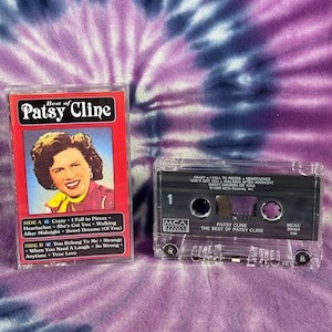 May include: A cassette tape of Patsy Cline's "The Best of Patsy Cline" album. The cassette tape is in a clear plastic case with a black label. The label has the title of the album, the artist's name, and the record label's logo. The cassette tape is on a purple and blue tie-dye background.