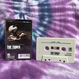 Könnte beinhalten: Eine Kassette und Hülle für das Album "The Tubes: Remote Control". Die Hülle ist schwarz mit einem Roboter-Artwork. Die Kassette ist weiß mit dem Albumtitel und Songtiteln bedruckt. Der Hintergrund ist ein Batikmuster in Lila und Weiß.