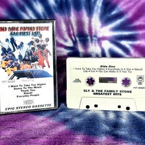 May include: A white cassette tape with the text "Sly & The Family Stone Greatest Hits" and "Side One" printed on it. The cassette tape has a black and white photo of the band on the front. The text on the cassette tape also includes "I Want To Take You Higher", "Everybody Is A Star", "Stand!" and "Life Fun You Can Make It If You Try".