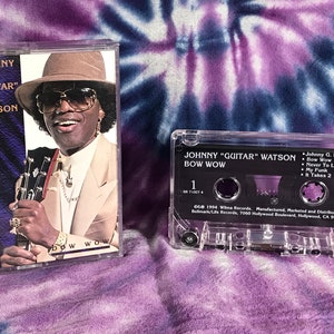 May include: A cassette tape with a black case and a clear plastic shell. The tape is labeled "Johnny "Guitar" Watson Bow Wow" and features a photo of Johnny Watson on the front. The back of the tape lists the songs on the cassette: "Johnny G. Is Back", "Bow Wow", "Never To Late", "My Funk", and "It Takes 2".