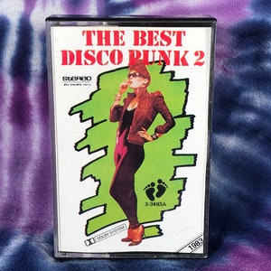 May include: A cassette tape with a green and white background featuring a woman in a black jumpsuit and a brown jacket. The text on the cassette tape reads "The Best Disco Punk 2" and "S-3493A".