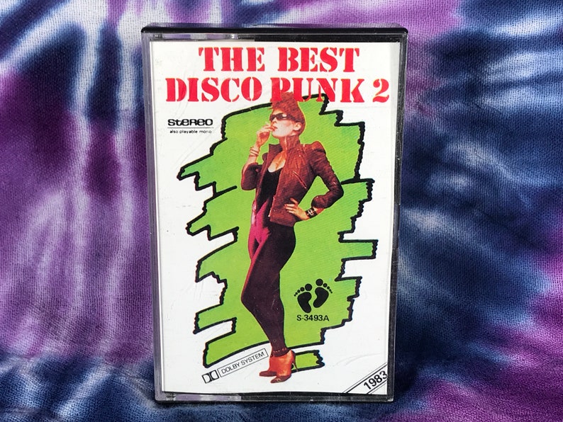 May include: A cassette tape with a green and white background featuring a woman in a black jumpsuit and a brown jacket. The text on the cassette tape reads "The Best Disco Punk 2" and "S-3493A".