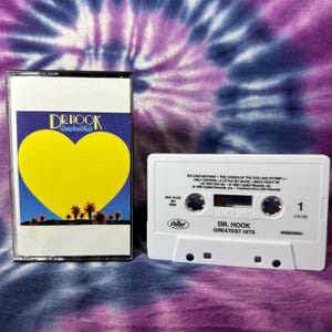 Puede incluir: Un casete de Dr. Hook Greatest Hits y su caja de plástico. La caja presenta un gran corazón amarillo sobre un cielo azul con palmeras. La carcasa blanca del casete muestra la lista de canciones, incluyendo Sylvia's Mother y The Cover of the Rolling Stone, con el logotipo de Capitol Records.