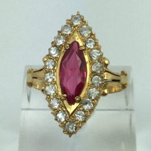 Puede incluir: Un anillo de oro con una gran piedra roja de forma marquesa rodeada de piedras pequeñas y transparentes.