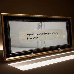 Puede incluir: Una obra de arte rectangular enmarcada con un borde negro y dorado. En el interior, se muestra un trozo de papel con texto hebreo. El marco está sostenido por dos soportes de plástico transparente. El fondo tiene un tono cálido.