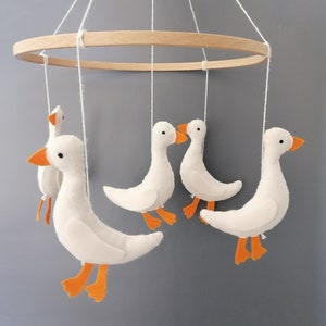 May include: A white wooden mobile with five felt geese hanging from it. The geese have orange beaks and feet. The mobile is hanging from a white ceiling.
