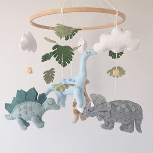 May include: A baby mobile featuring a light blue dinosaur, a green dinosaur, a grey dinosaur, and white clouds. The mobile is crafted from felt and has a wooden ring.