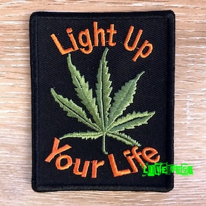 May include: Black rectangular patch featuring the phrase "Light Up Your Life" in orange. A green cannabis leaf is embroidered in the centre. This patch is designed to be sewn or ironed onto clothing or accessories.