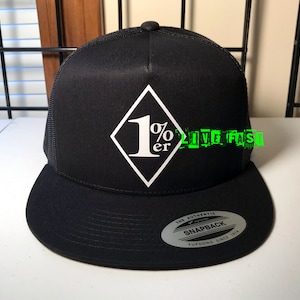 Op de afbeelding: Zwarte truckercap met een wit diamantlogo met de tekst "1%er". De pet heeft een zwart mesh achterpaneel en een zwarte drukknopsluiting. De pet is gemaakt door Yupoong en is een authentieke snapback.
