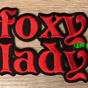 Puede incluir: Parche de hierro bordado en rojo y negro con el texto "Foxy Lady" en una fuente estilizada.