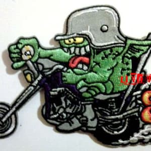 May include: Embroidered patch depicting a green monster on a motorcycle. The monster wears a grey helmet and has a red tongue. The motorcycle is black and purple, with flames and smoke. The patch is on a white background.